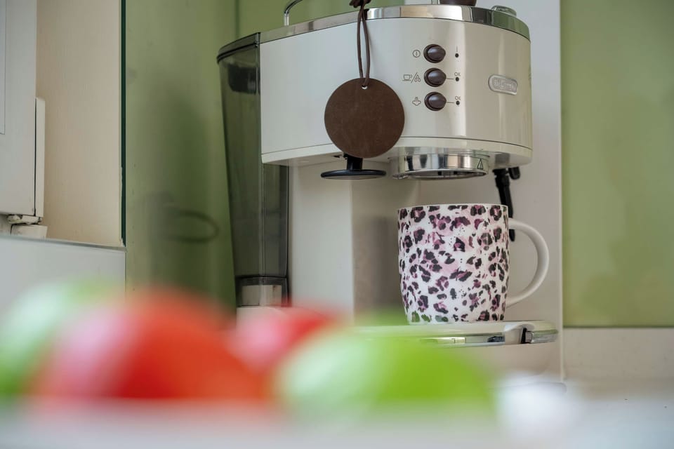 A silver coffee machine sits on a kitchen counter with a patterned mug underneath it. The background has green walls and kitchen cabinets, with blurred red and green objects in the foreground.