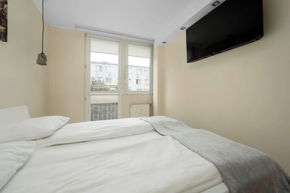 A bright bedroom with a double bed covered in white bedding and a light-gray throw. A wall-mounted television hangs above the bed area, and a large window behind the bed brings in daylight.