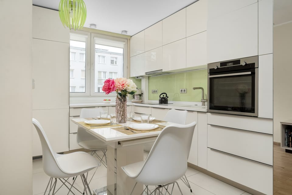 A modern white kitchen with sleek cabinets, a built-in microwave, and a light countertop. A small dining table with white chairs sits in front of the window, with a vase of flowers adding color.