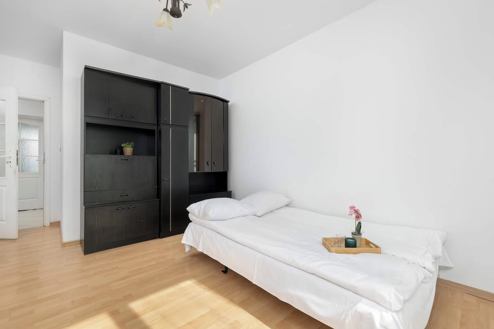 A bedroom area includes a white sofa bed positioned next to a large black wardrobe. Sunlight illuminates the white walls and wooden floor.