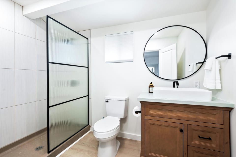 Primary ensuite, step into your bright, modern bathroom where clean lines meet warm wood tones. A spacious walk-in shower with elegant frosted glass awaits, while a sleek vessel sink and oversized circular mirror complete your private retreat.