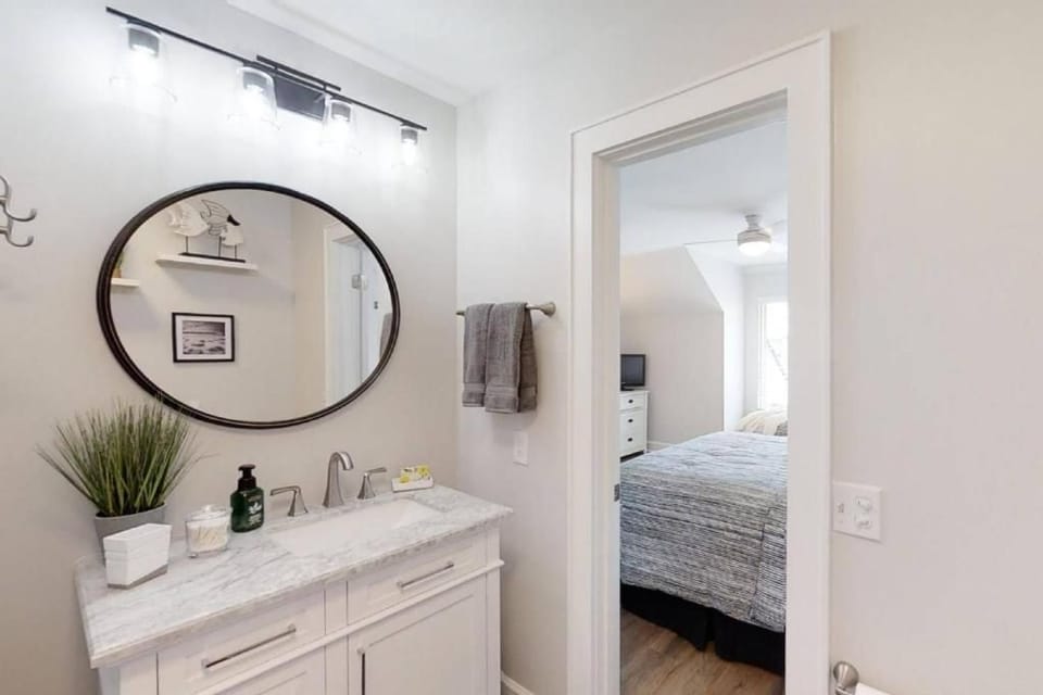 Glowing bulbs illuminate this tidy ensuite, where a round mirror reflects a calm and organized space. Stone counters and silver fixtures provide a clean finish, leading your eye through the doorway into a sunlit, peaceful bedroom.