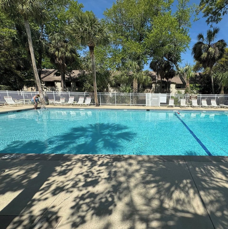 Azure waters shimmer beneath towering palms and a bright midday sky. Soft dappled shadows dance across the warm concrete deck, offering a quiet invitation to spend a slow, sun-drenched afternoon by the pool.