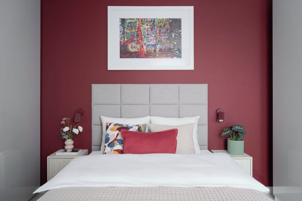 A modern bedroom with a large double bed, red accent cushions, and a burgundy wall behind the headboard. Small bedside tables with lamps and decorative plants create a welcoming atmosphere.