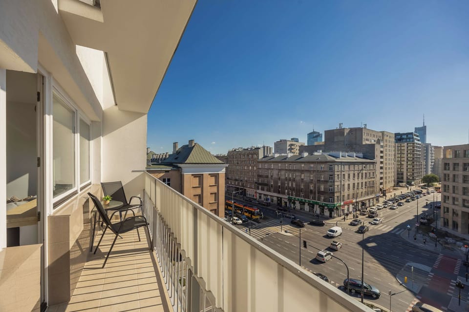 A spacious balcony with outdoor seating overlooking the surrounding city. The view includes nearby buildings, streets, and a clear blue sky.