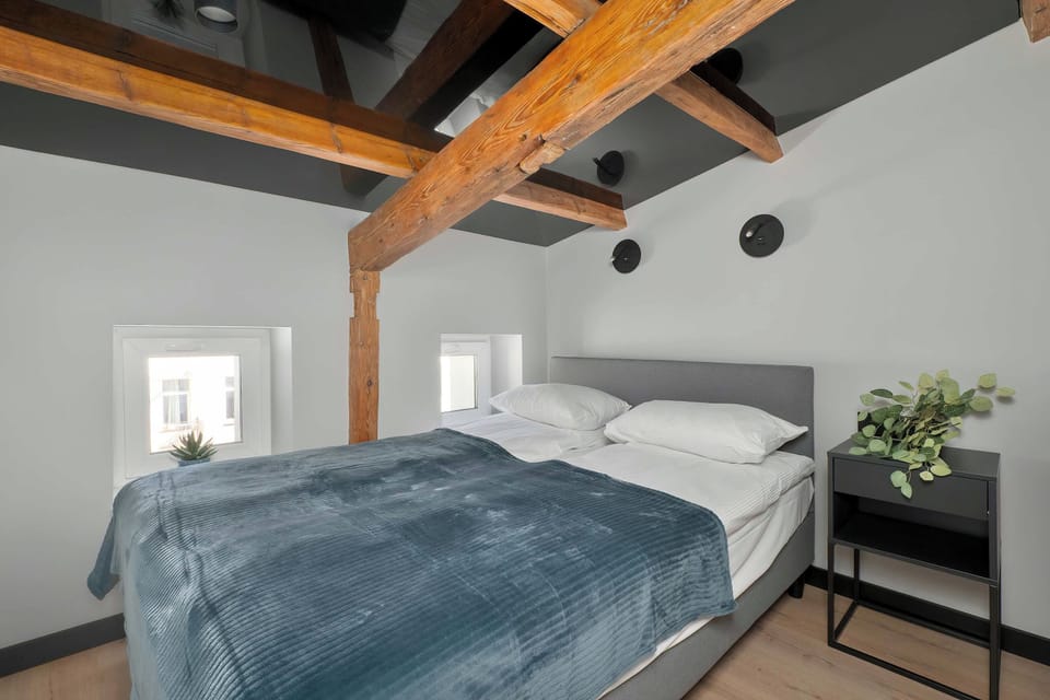 A bedroom furnished with a large bed covered in a dark blue-gray blanket, framed by exposed wooden beams and white walls.