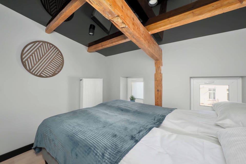 A bedroom with a neatly made bed covered in a blue-gray blanket, a round wall decoration, and warm wooden ceiling beams.