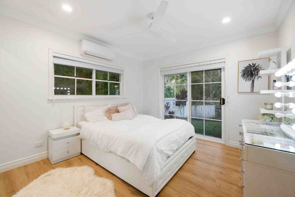 Light-filled guest bedroom styled with white bedding, a vanity mirror station, and large windows looking out to greenery.