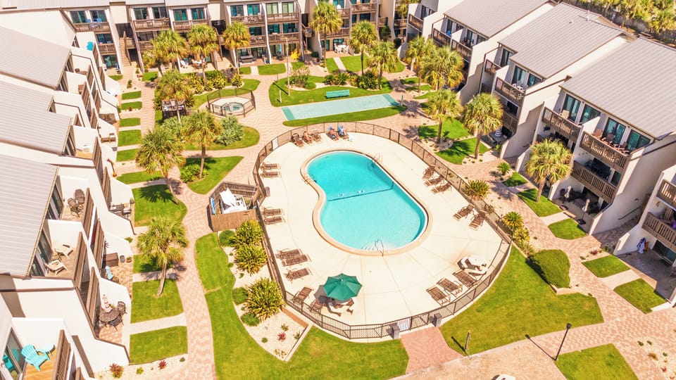 A bird's-eye view showcases the property's well-maintained pool area with surrounding sun loungers, lush green lawns, palm trees, and multi-story residential buildings arranged around a central courtyard.