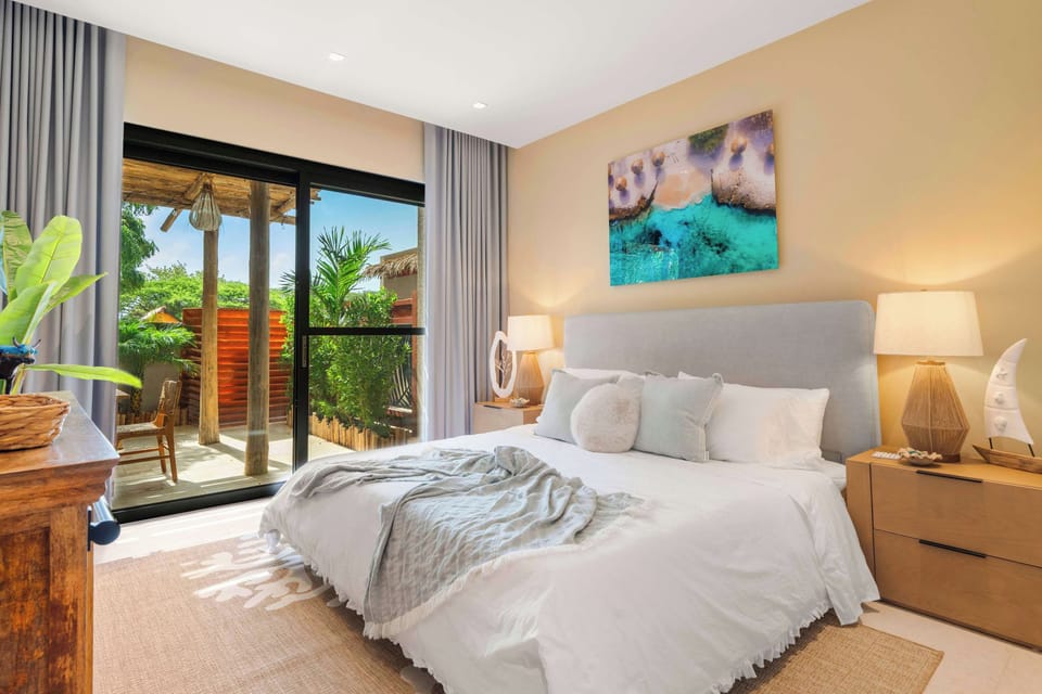 A bright, relaxing bedroom with soft neutral finishes, tropical artwork, and doors opening directly to the private patio.