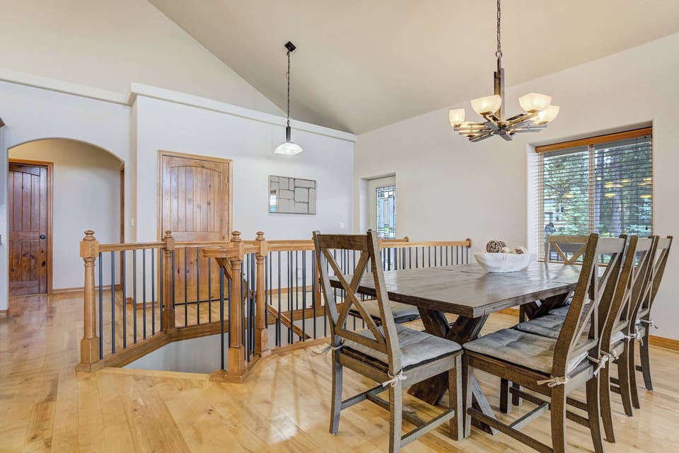 Just off the kitchen, a farmhouse trestle table with cross-back chairs sits beneath a wrought iron chandelier. A nearby window lets in soft daylight, making it an inviting spot for family dinners, morning coffee, or a puzzle.