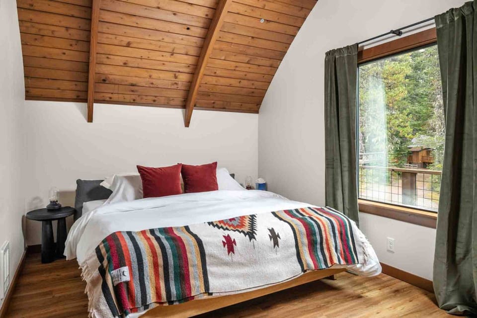 Bedroom view under a sloped wooden ceiling with white bedding, vibrant red pillows, and forest window views.