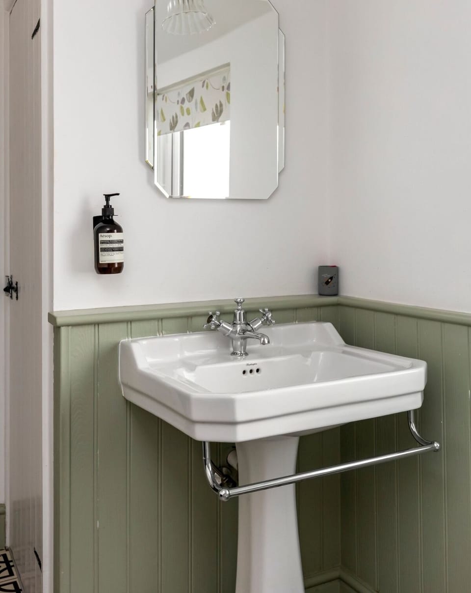 A charming, traditional-style bathroom with antique-style basin. Thoughtful details include hand soap and mirrors with their own integrated lighting, combining classic character with practical, modern touches for a comfortable, well-considered space.