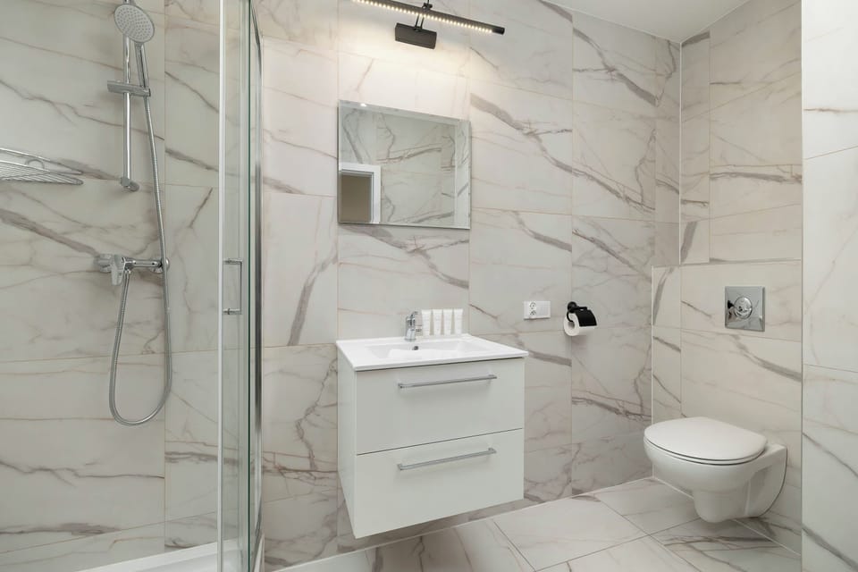 A bright bathroom with marble-patterned wall tiles, a white vanity with a sink, and a toilet. A glass-enclosed shower area is visible on the right side.