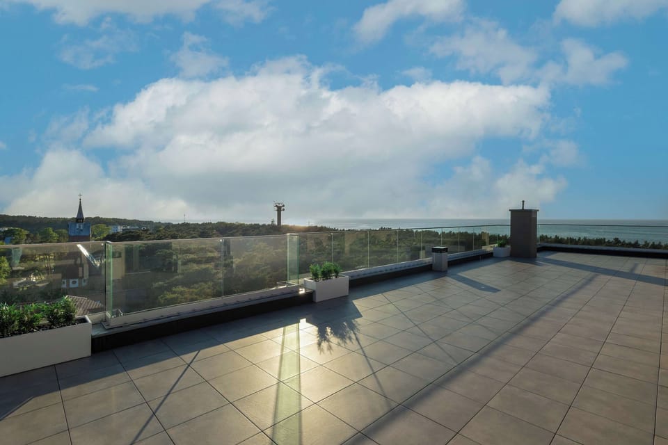 An expansive rooftop terrace offers panoramic views of the surrounding green area under a cloudy blue sky. The large tiled space is bordered by sleek glass railings and built-in planters.