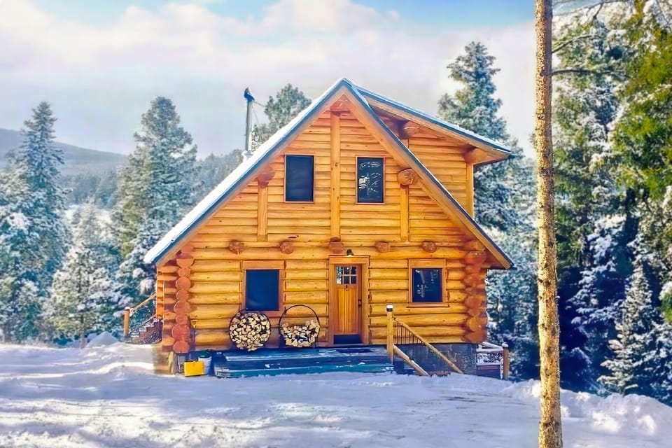 Snowy two-story log cabin with porch, firewood, and pine forest views