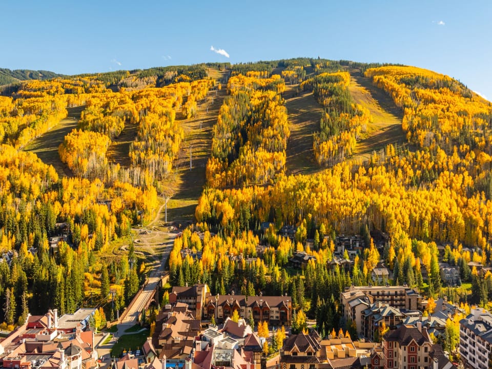 A stunning mountain resort blanketed in golden autumn foliage, with ski runs cutting through vibrant aspen groves and charming alpine village buildings nestled at the base.
