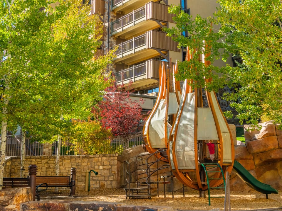 The playground features unique wooden structures and slides nestled among vibrant autumn foliage, perfect for family fun just steps from your rental.