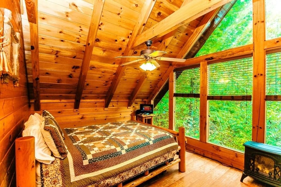 Queen sized bed in the loft area with a wall of windows overlooking the woods