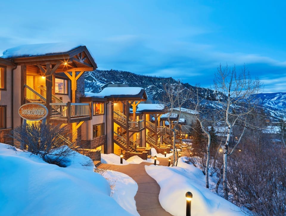 A mountain resort property nestled in snow-covered peaks, featuring alpine architecture with timber framing and warm exterior lighting against the winter twilight sky.