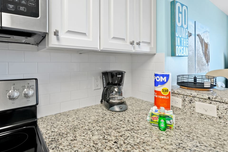 Your kitchen features granite countertops, white cabinetry, and a coffee maker ready for your morning brew. Coastal-themed art adds charm to the space.