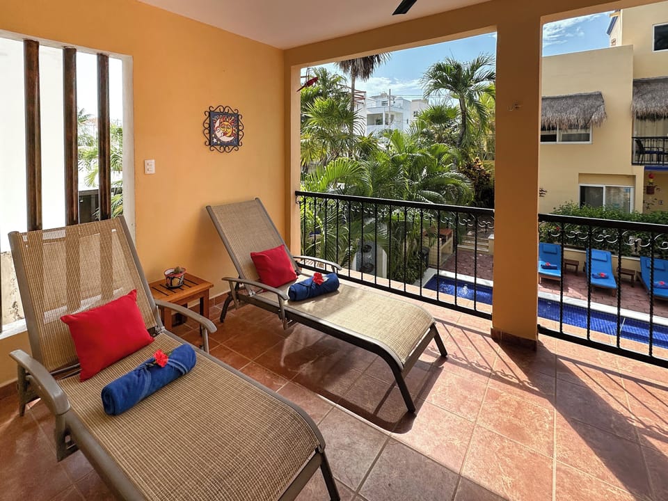 A2-10-2026 Balcony Lounge Chairs - Private balcony at Abbey del Sol A2 Unit 10, a Puerto Morelos vacation rental, featuring lounge chairs, colorful cushions, shade, tropical plants, and a view over the pool and courtyard.