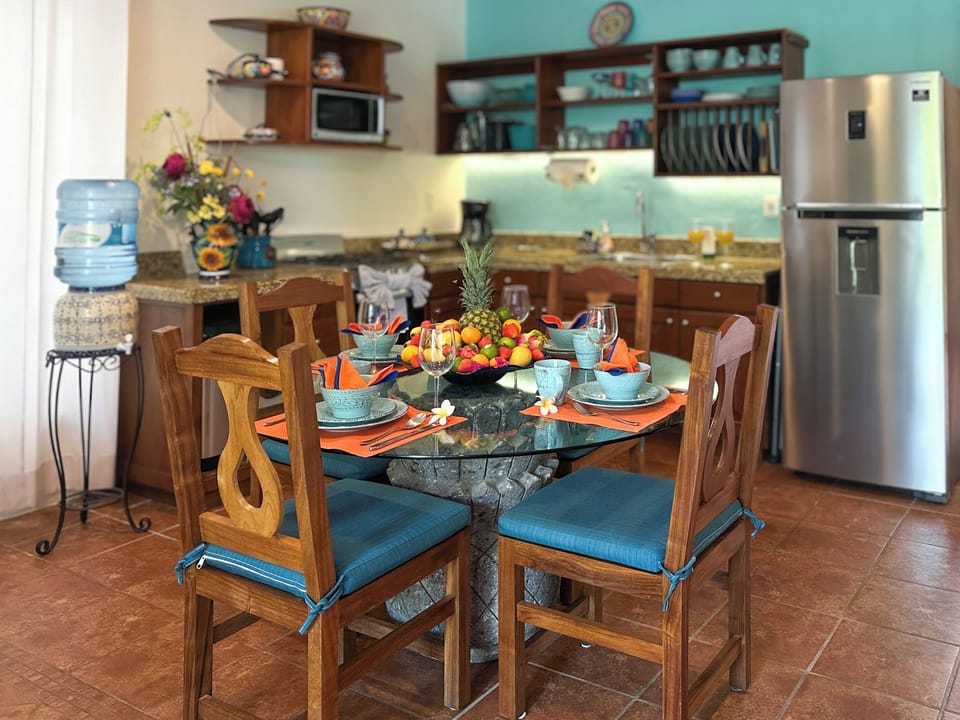 A2-10-2026 Kitchen and Dining Area - Kitchen and dining area inside Abbey del Sol A2 Unit 10, a Puerto Morelos vacation rental, featuring a dining table with colorful place settings, wood chairs, stainless refrigerator, water dispenser, and a well-equipped kitchen for a comfortable stay.