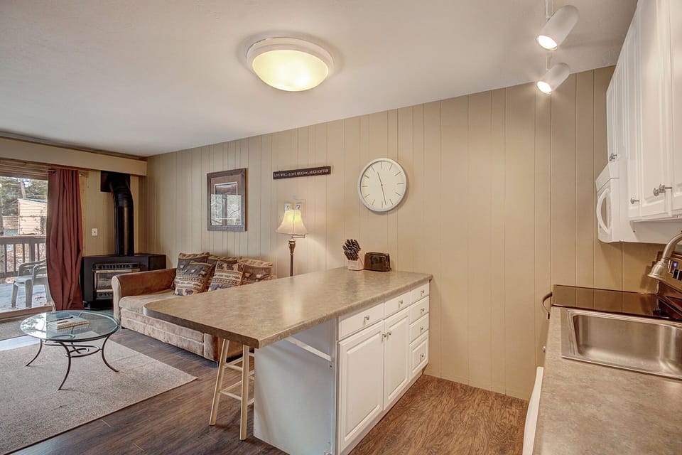 A cozy living room and kitchen area features a small sofa, wall clock, wood paneling, and modern lighting fixtures. A glass coffee table and a wood-burning stove add to the room's comfort and functionality.