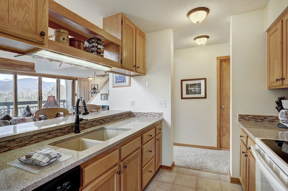 A kitchen with wooden cabinets, a white countertop with a double sink, and a view of the living room and mountains in the background. The hallway leads to other rooms.