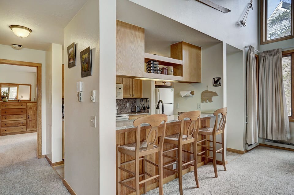 Kitchen area with a breakfast bar featuring three wooden stools, adjacent to a hallway leading to a bedroom. The kitchen has wooden cabinets, a refrigerator, and is accented by small decor items on the wall.