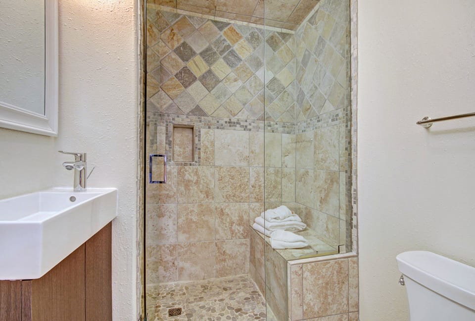A modern bathroom features a glass-door shower with tiled walls and a built-in bench, neatly folded towels on the bench, a white sink with a mirror above, and a toilet to the right.