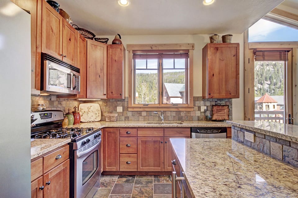 A kitchen with wooden cabinets, a stainless steel stove and microwave, granite countertops, a window with a scenic view, and stone tile flooring.