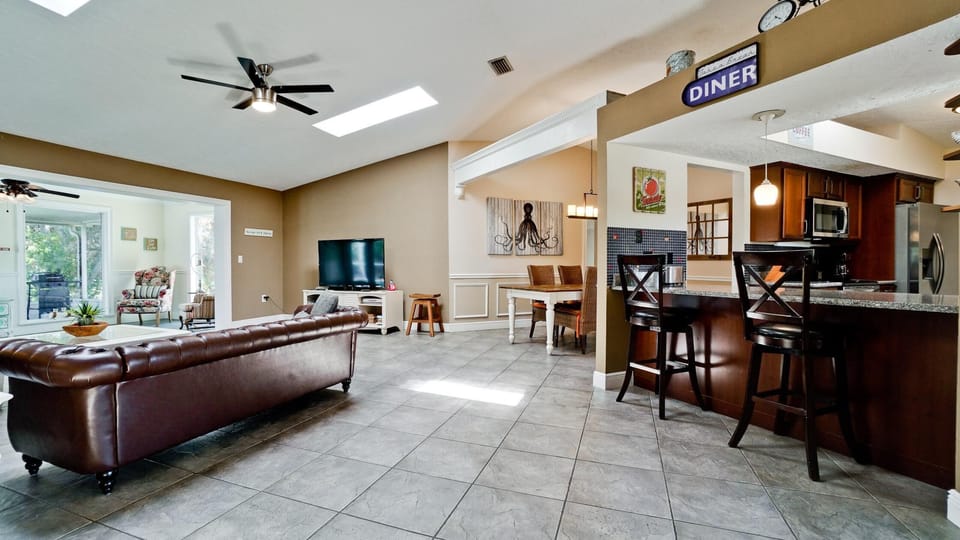 Open living area with large brown leather sofa, tiled floors, skylight, and adjacent kitchen bar with dark wood stools.