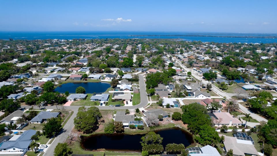Aerial view of a quiet residential neighborhood with lakes, single-story homes, green trees, and distant waterfront.