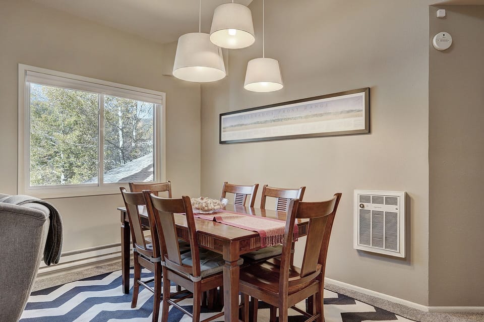 Dining area with a wooden table and six chairs, pendant lights above the table, a framed picture on the wall, and a window with a view of trees outside. A chevron-patterned rug is on the floor.