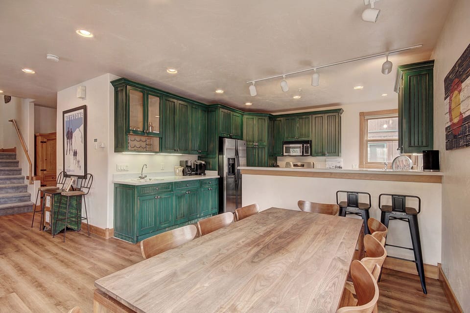 A spacious kitchen and dining area featuring green cabinets, a wooden dining table with chairs, bar stools at a counter, and modern appliances. The room has wooden flooring and recessed lighting.