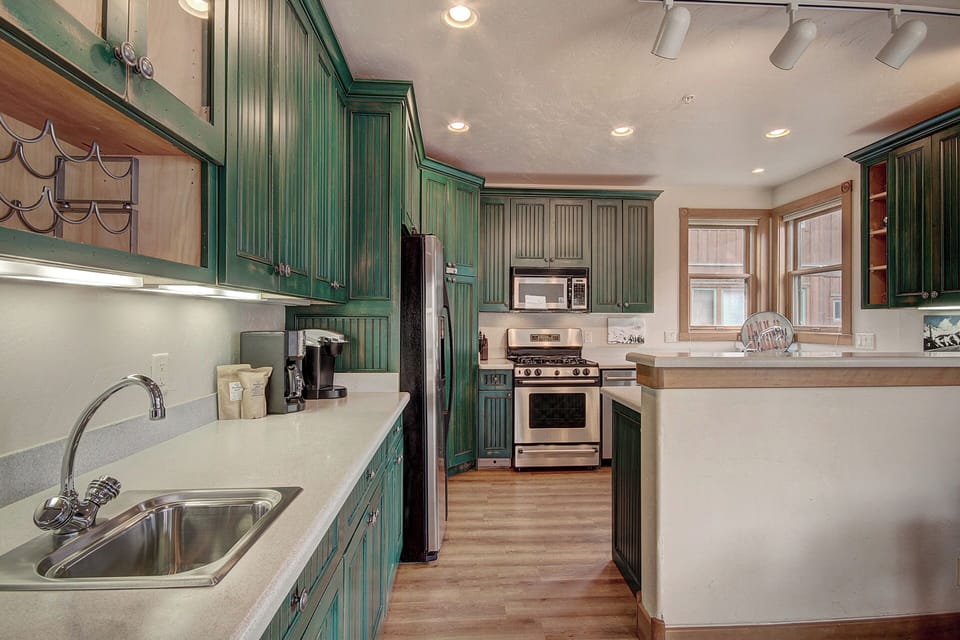 A modern kitchen with green cabinets, a stainless steel stove, microwave, and refrigerator. There is a sink with a faucet, a coffee maker, and natural light coming in from a window.