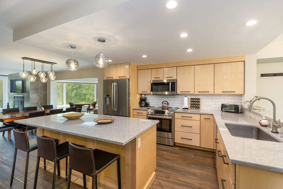 A modern kitchen with light wood cabinets, stainless steel appliances, a large island with seating for three, and a view into a dining area with a table and chairs next to large windows.
