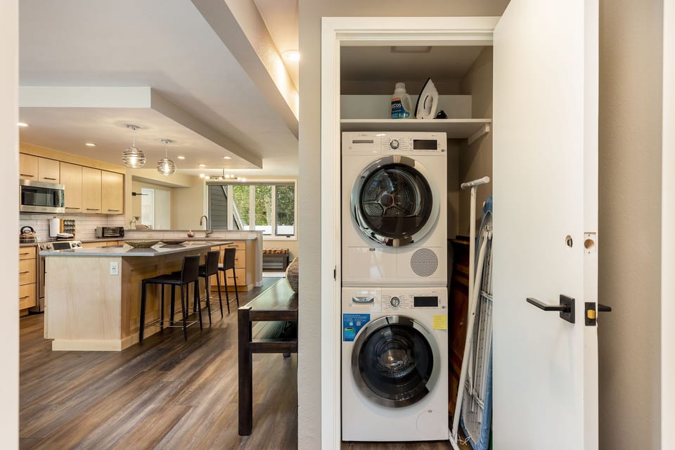 A modern kitchen with light wood cabinets, stainless steel appliances, and a center island is adjacent to a laundry closet with a stacked washer and dryer. An ironing board is visible next to the closet door.