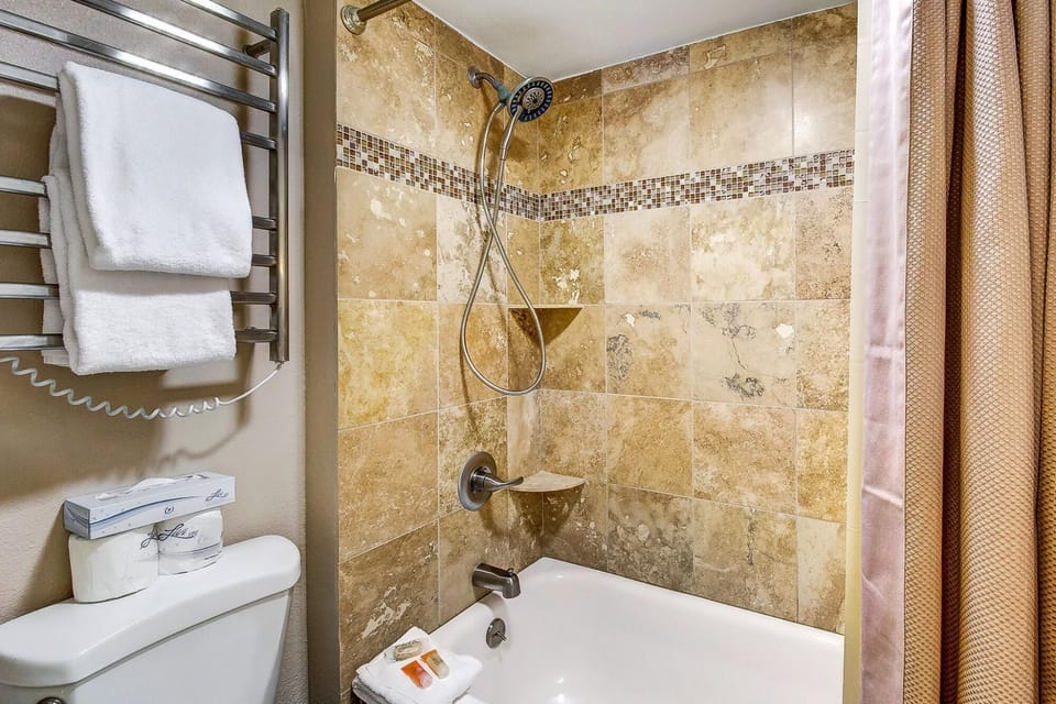 A bathroom featuring a bathtub with a tile backsplash, a showerhead, a towel rack with towels, a toilet, and a beige shower curtain.