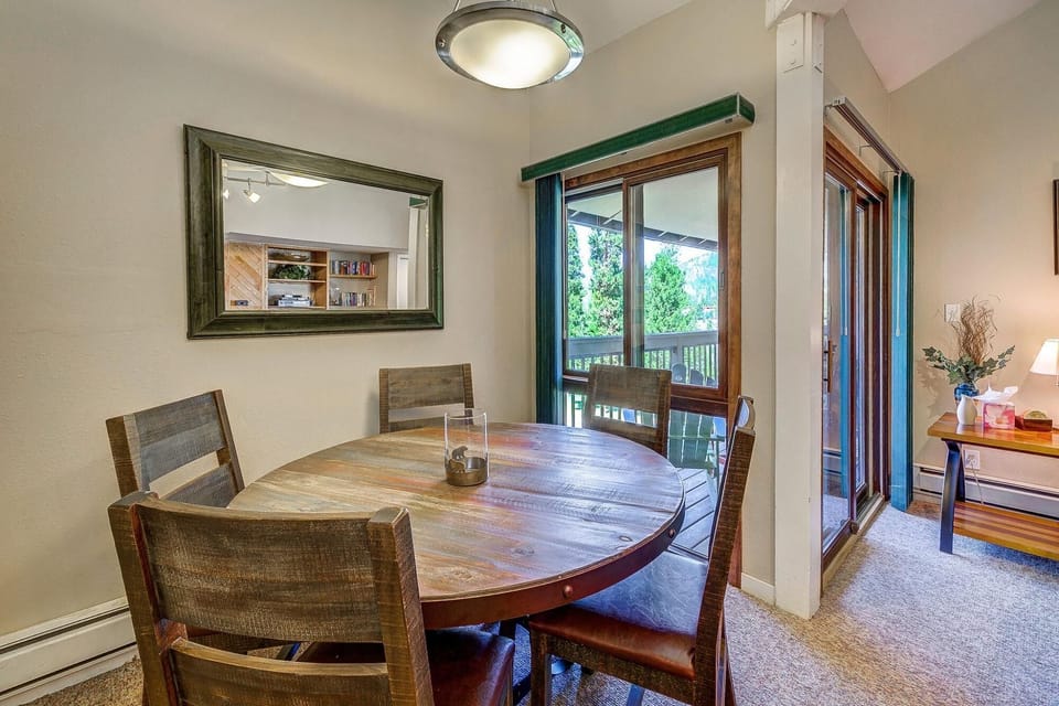 A dining area with a round wooden table and four chairs, illuminated by a ceiling light. A sliding glass door opens to a balcony, with a window and mirror on adjacent walls.