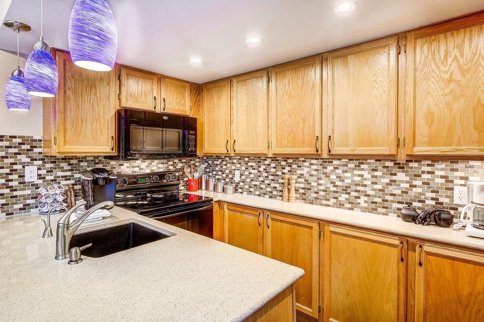 A modern kitchen with wooden cabinets, a mosaic tile backsplash, black appliances, a beige countertop with a sink, and blue pendant lights.