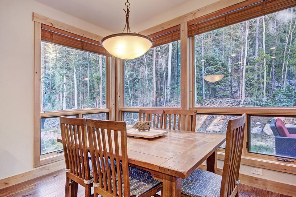A dining area with a wooden table and six chairs, large windows showcasing a forested outdoor view, and a hanging light fixture above the table.