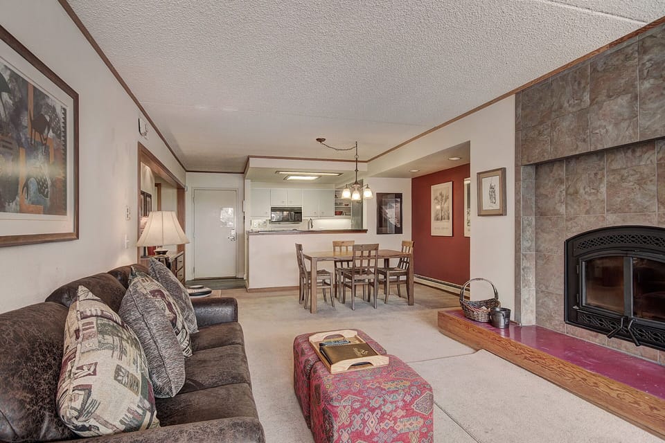A living room with a brown couch, patterned cushions, a red ottoman, and a fireplace. A dining table with chairs is seen near the open kitchen area in the background. Various framed artworks adorn the walls.