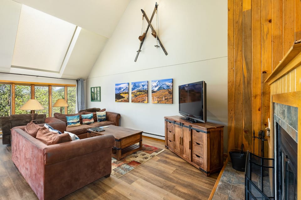 A living room with a brown sectional sofa, a wooden coffee table, a flat-screen TV on a wooden stand, and a fireplace. Wall decor includes crisscrossed skis and three landscape pictures.