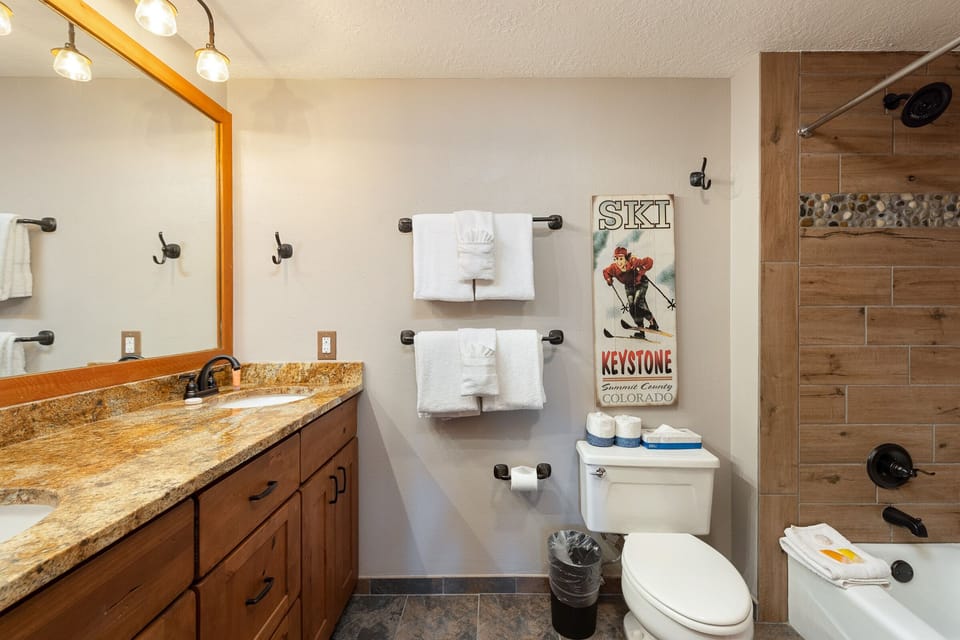Bathroom with a double vanity, granite countertop, wooden cabinets, towel racks with white towels, toilet, and a bathtub. Wall has a vintage ski poster.