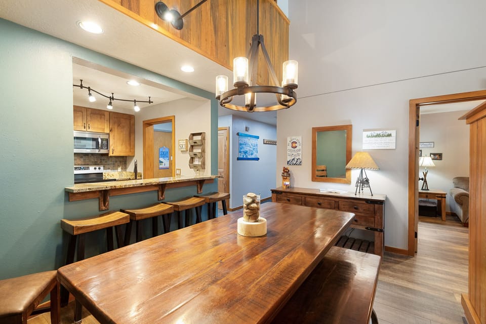 A cozy, rustic dining area with a wooden table and benches, a chandelier, a kitchen with a bar and stools, and light brown walls adorned with decor accents.