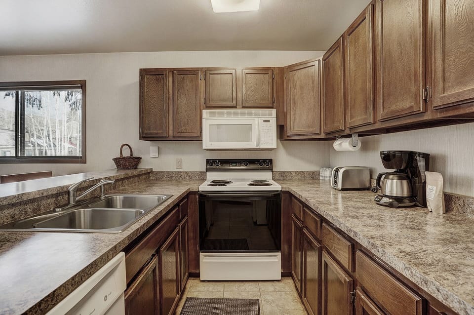 A kitchen with wood cabinets, a double sink, an electric stove, a microwave, a coffee maker, a toaster, and a window. The countertop and backsplash feature a stone-like pattern.