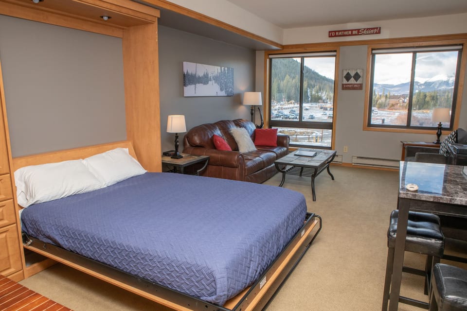 A studio apartment features a bed with a blue quilt, a brown leather sofa, two windows providing a mountain view, and a wall-mounted TV. The decor is light and modern.