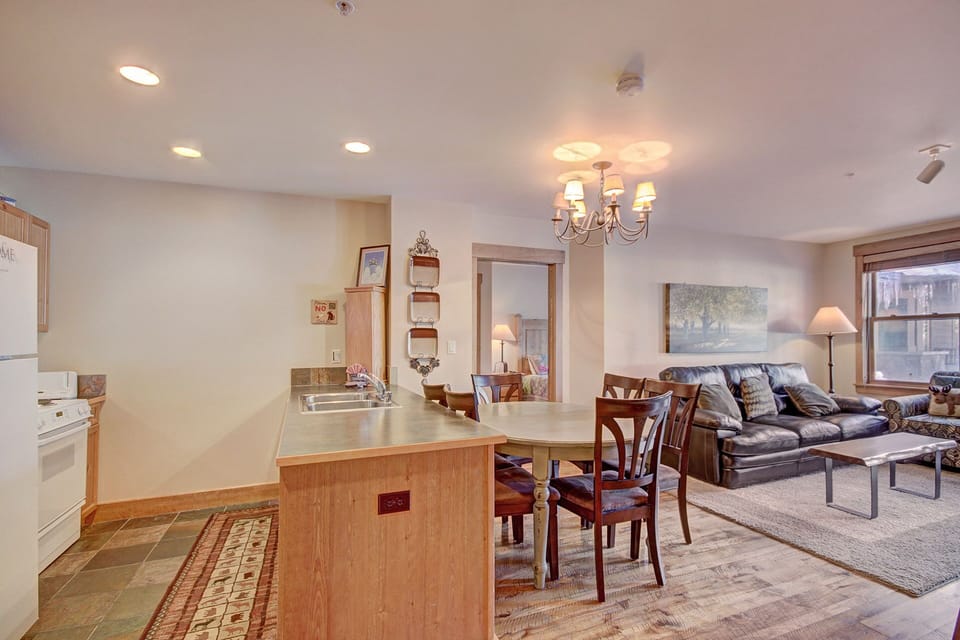 Open-concept living and dining area with a view of the kitchen. Features wooden dining table and chairs, leather sofa, chandelier, and wall art, with ample natural light from windows.